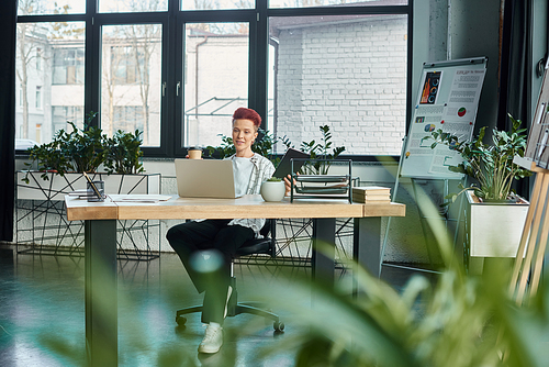 stylish non-binary person with coffee to go and notebook sitting near laptop at workplace in office