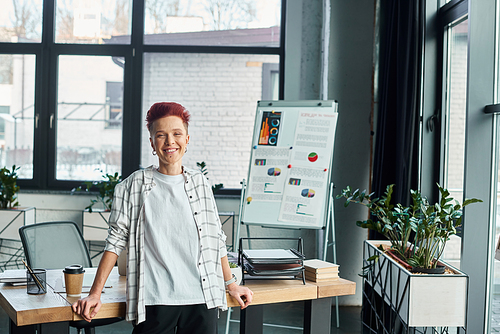 happy successful bigender person standing at workplace in modern office and looking at camera