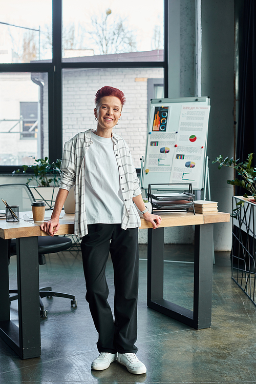 happy successful bigender person standing at workplace in modern office and looking at camera