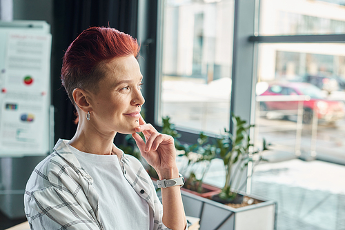 pensive and smiling non-binary manager in casual attire standing and looking away in modern office