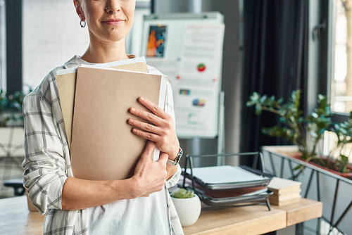 cropped view of non-binary manager holding folders with documents while standing in modern office