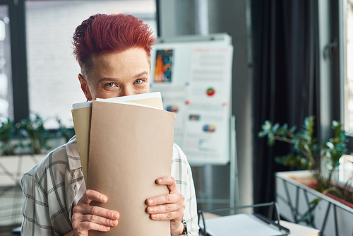 cheerful bigender person with short hair obscuring face with folders and looking at camera in office