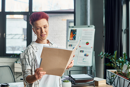 stylish bigender person in casual attire looking in folder with documents in contemporary office