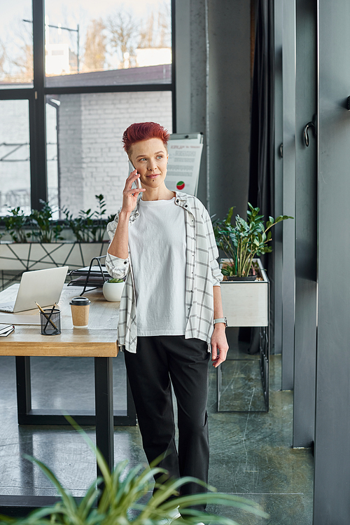 stylish bigender manager in casual attire standing and talking on smartphone in modern office