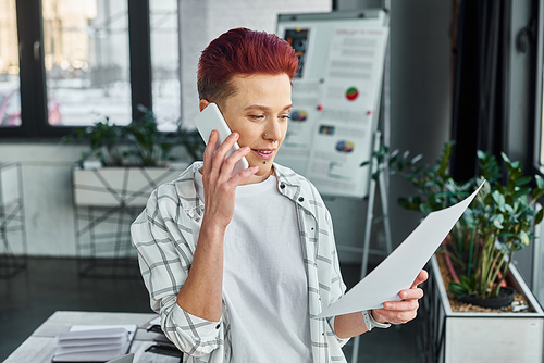 bigender manager in casual attire looking at document and talking on smartphone in modern office