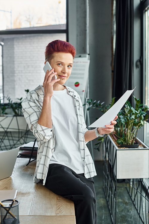 queer person in casual attire sitting on work desk with document and talking on smartphone in office