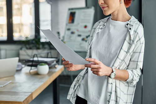 cropped view of non-binary person in casual attire standing with paper documents in modern office