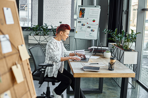 queer person in casual attire typing on laptop while sitting near documents on work desk in office