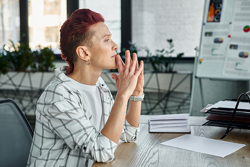 side view of thoughtful non-binary manager with hands near face sitting at workplace in office