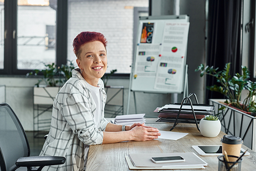 joyful queer manager sitting at workplace near smartphone and digital tablet and looking at camera