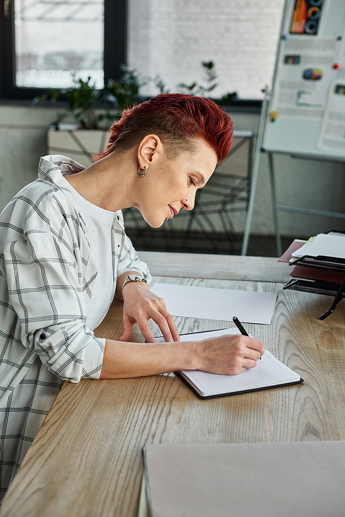 concentrated stylish bigender person sitting at workplace in modern office and writing in notebook