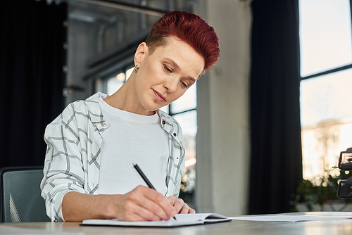 concentrated stylish bigender person sitting at workplace in modern office and writing in notebook