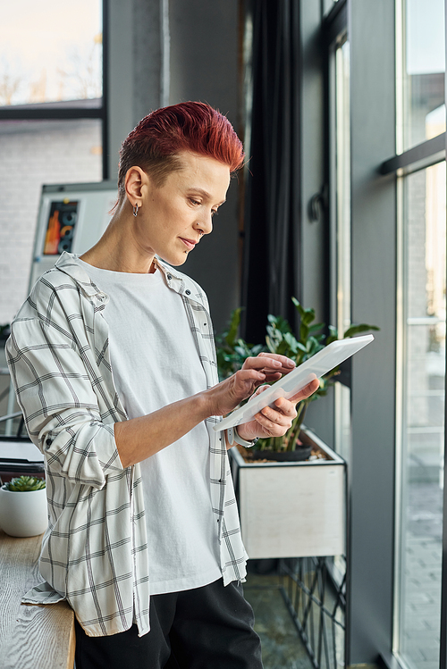 stylish bigender manager in casual attire standing in modern office and using digital tablet