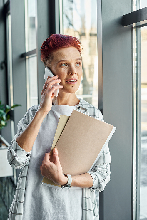 stylish bigender manager standing near window with documents and talking on smartphone in office