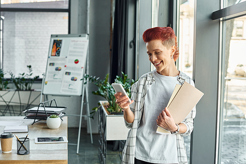 happy queer person in casual attire standing with documents in office and messaging on smartphone