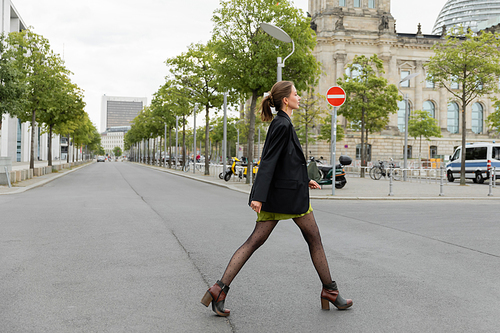 Side view of fashionable young woman in dress and black jacket walking on road in Berlin, Germany