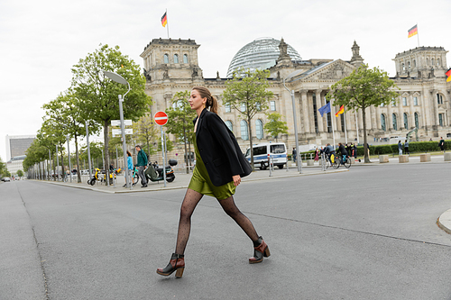 Full length of young and fair haired woman in silk dress, jacket and boots walking on road in Berlin