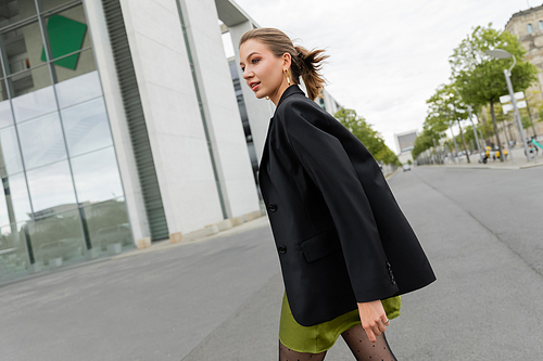 Smiling young and fair haired woman in black blazer, silk dress looking away while walking in Berlin