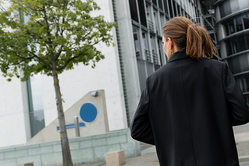 Back view of stylish and fair haired young woman in black jacket walking in Berlin, Germany