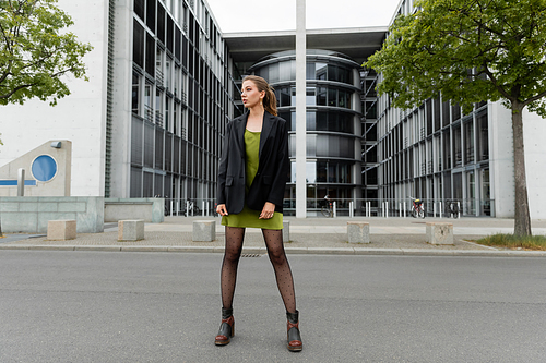 Full length of trendy young woman in boots and stylish blazer looking away  on street in Berlin