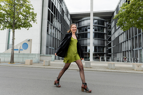 cheerful and fashionable young woman in blazer and green silk dress in Berlin, Germany