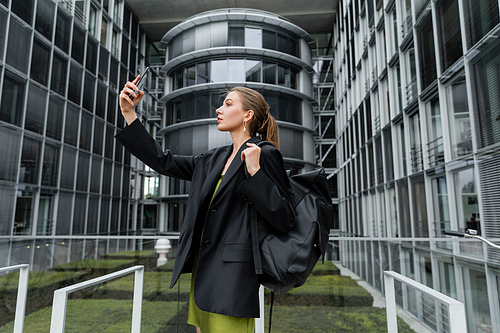 Side view of fashionable young woman in jacket holding backpack and taking selfie on smartphone