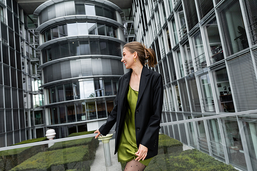 Side view of cheerful young fair haired woman in black jacket and silk dress standing in Berlin