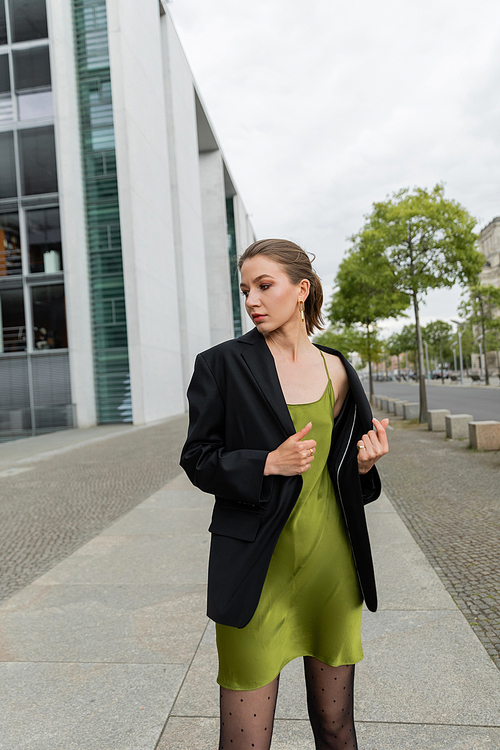 Stylish young woman in black blazer and silk dress looking away while standing in Berlin, Germany