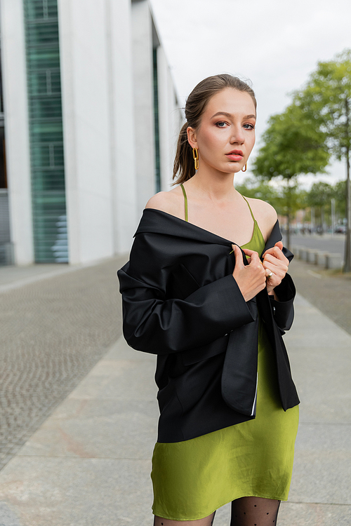 Young fair haired woman in stylish silk dress posing in black blazer and looking at camera in Berlin