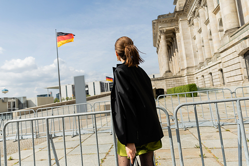 Back view of fair haired woman in jacket and dress standing near blurred Reichstag Building