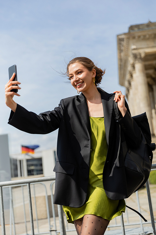 Cheerful woman in jacket and silk dress holding backpack and taking selfie  in Berlin, Germany