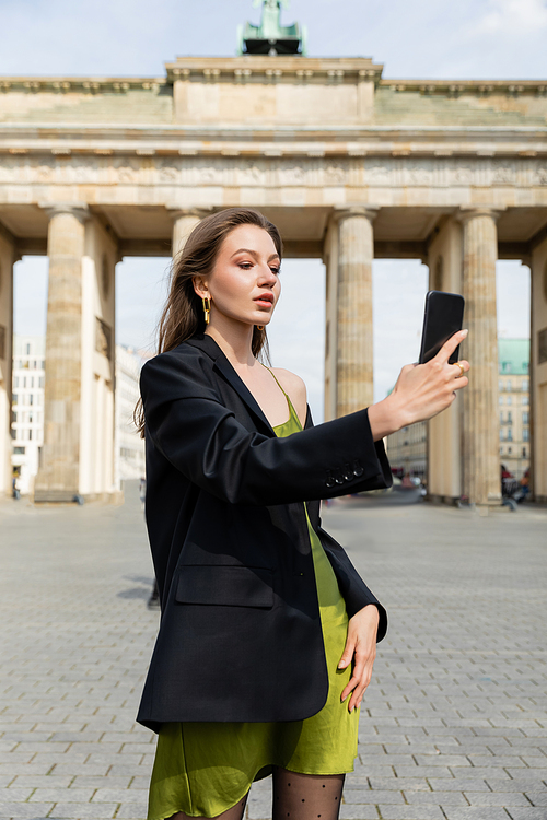 elegant fashionable woman in jacket and silk dress taking selfie near Brandenburg Gate in Berlin