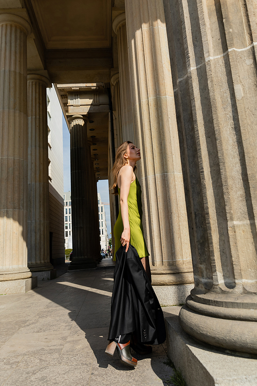 Elegant young woman in silk dress holding black jacket and standing in Berlin, Germany