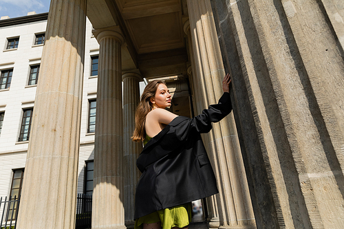 trendy young woman in silk dress holding black jacket and standing in Berlin, Germany