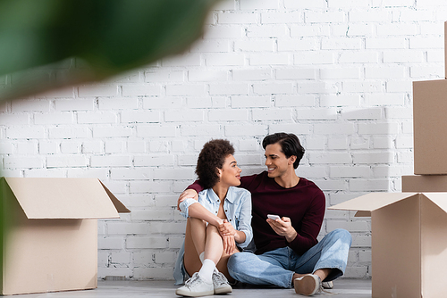 happy multiethnic couple sitting on floor and looking at each other near carton boxes