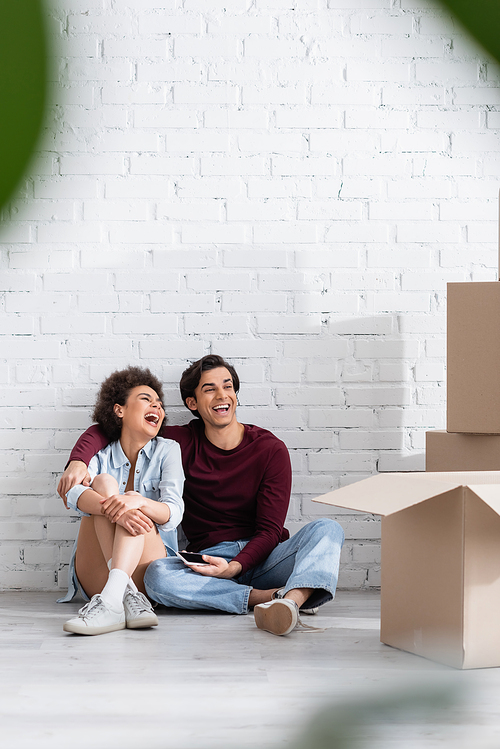 happy multiethnic couple sitting on floor near carton boxes