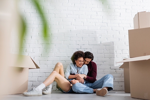 happy multiethnic couple sitting on floor near carton boxes