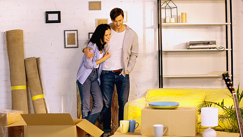 happy man standing with hand in pocket and hugging cheerful girlfriend in new home