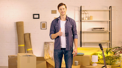 happy young man showing thumb up near cardboard boxes in new home