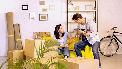 cheerful man with acoustic guitar near happy woman on couch in new home