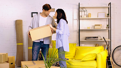 happy young man holding box near cheerful girlfriend in living room