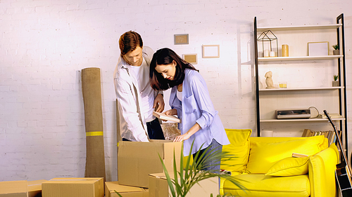 young woman holding book while looking at box and unpacking near boyfriend