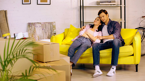 Smiling couple talking and resting on couch near boxes in new home