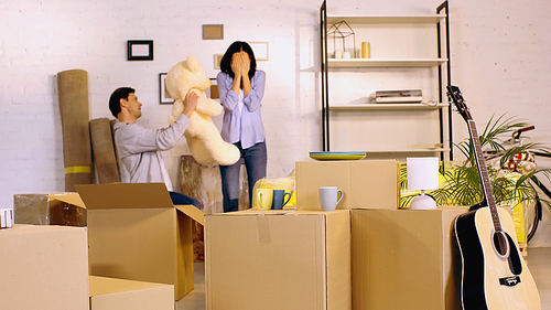 man holding teddy bear near girlfriend covering eyes in living room