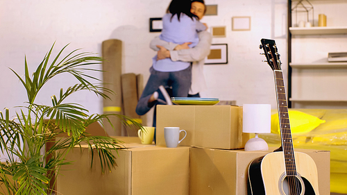 acoustic guitar, plant and carton boxes near blurred couple hugging on background