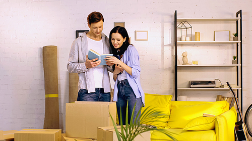 cheerful young couple reading book near boxes in living room