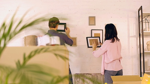 back view of couple hanging photo frames on wall near boxes in new home