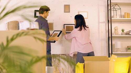 Couple hanging photo frames on wall near boxes in new home