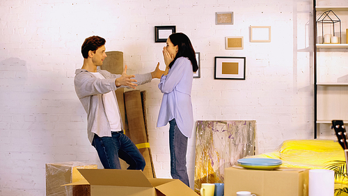 excited woman covering mouth and looking at boyfriend with outstretched hands in new home