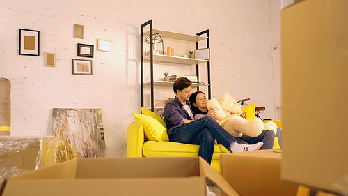 happy couple chilling on sofa with teddy bear near boxes in new home